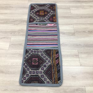 May include: A long, rectangular rug with three panels. The top panel is a dark purple and black geometric pattern. The middle panel is a multi-colored striped pattern. The bottom panel is a dark purple and black geometric pattern.