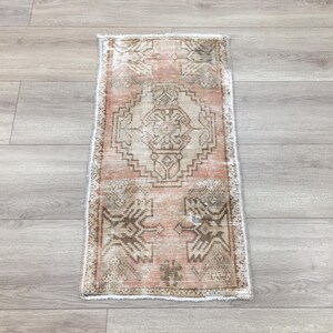 May include: A rectangular area rug featuring a faded peach and beige color palette with intricate geometric patterns. The design includes a central diamond motif surrounded by symmetrical tribal-inspired shapes and a fringed edge on both ends.