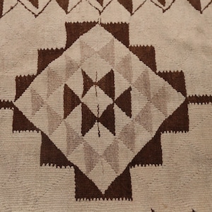 May include: A close-up of a woven rug with a geometric pattern in shades of brown and white. The pattern features a central diamond shape surrounded by smaller triangles and squares.