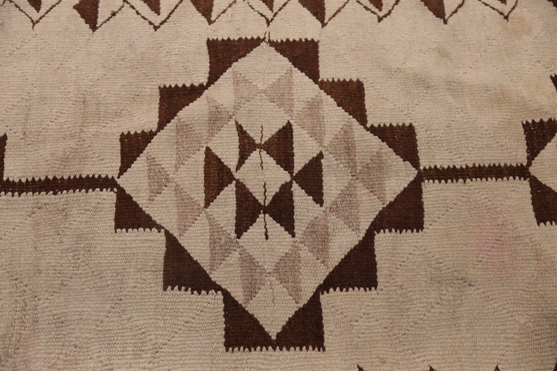 May include: A close-up of a woven rug with a geometric pattern in shades of brown and white. The pattern features a central diamond shape surrounded by smaller triangles and squares.