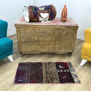 May include: A rectangular wooden chest with intricate floral carvings on the front panel, topped with a cylindrical duffel bag featuring a colourful patterned textile and brown leather accents, alongside a small terracotta lantern and a rug.
