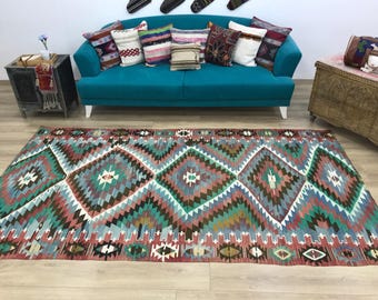 May include: A rectangular area rug featuring a repeating geometric diamond pattern in shades of teal, brown, pink, and cream. The design includes intricate zigzag borders and small symmetrical motifs throughout the woven fabric surface.