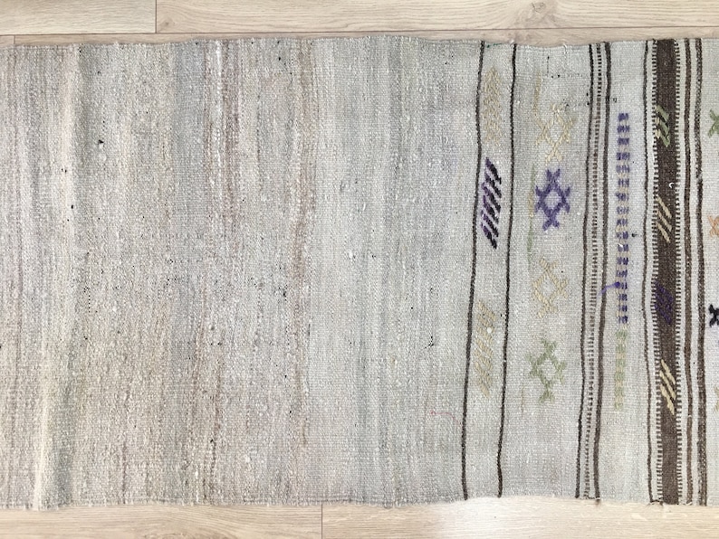 May include: A light gray rug with a woven pattern in brown, white, and purple. The rug has a fringe border.