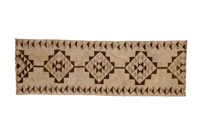 May include: A long, rectangular rug with a beige background and a brown geometric pattern. The pattern features repeating diamond shapes with smaller triangles inside.