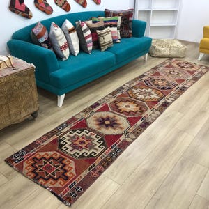 May include: A long, rectangular runner rug featuring a repeating geometric pattern in shades of red, orange, brown, and cream. The design consists of interconnected octagonal and star-shaped motifs with intricate borders, creating a textured appearance.