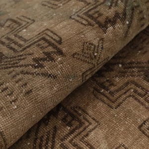 May include: A folded area rug featuring a muted brown and tan color palette with dark brown geometric patterns. The textile displays a woven texture with visible pile and intricate linear motifs that repeat across the surface of the fabric.