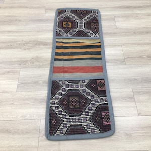 May include: A long, rectangular, gray fabric cushion with three different patterned panels. The top panel has a geometric design in shades of gray, white, and brown. The middle panel has horizontal stripes in shades of brown, white, and red. The bottom panel has a geometric design in shades of gray, white, and purple.