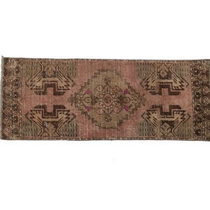 May include: A rectangular woven rug featuring a dusty pink central field with a large brown geometric diamond medallion. The design includes dark brown angular motifs on the sides and a decorative border with small floral patterns in brown and pink.