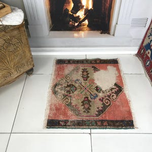 May include: A small, rectangular, distressed area rug featuring a central geometric medallion in muted shades of coral, sage green, and dark brown. The rug has a worn, faded patch on the right side, dark borders, and short white fringe on the ends.