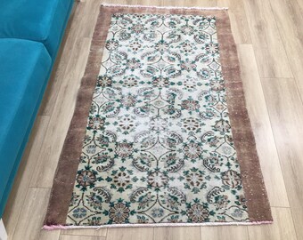 May include: A rectangular area rug featuring a central cream-colored field with a repeating floral and vine pattern in shades of teal, brown, and muted green. The rug is framed by a wide, solid brown border and finished with short fringe ends.