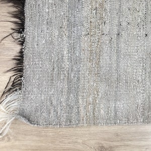 May include: A close-up of a gray and white woven rug with a fringe edge. The rug is made of natural fibers and has a rustic, hand-woven look.