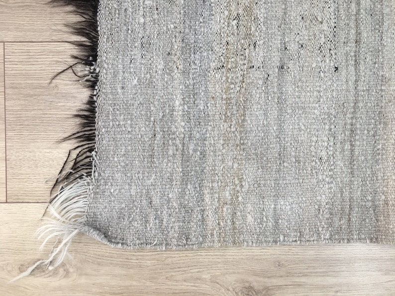 May include: A close-up of a gray and white woven rug with a fringe edge. The rug is made of natural fibers and has a rustic, hand-woven look.