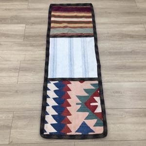 Puede incluir: Una alfombra larga y rectangular con tres secciones de diferentes patrones. La sección superior es un patrón de rayas marrones, blancas y rojas. La sección del medio es un patrón de rayas blancas y azules. La sección inferior es un patrón geométrico colorido con rojo, azul, verde y blanco.
