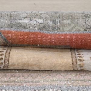 Può includere: Un tappeto tessile piegato con una composizione a strati e sezioni distinte. Lo strato superiore mostra un motivo geometrico grigio e bianco, mentre la piega centrale rivela una fascia arancione vibrante sopra una sezione beige con motivi marroni.