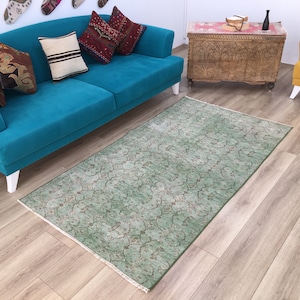 Op de afbeelding: Een groen tapijt met een patroon en een vervaagd ontwerp, geplaatst voor een turquoise blauwe bank met witte poten. De bank heeft verschillende kussens in verschillende kleuren en patronen.
