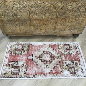 Puede incluir: Una alfombra rectangular con un patrón central de diamante en tonos terracota, crema y marrón oscuro. La alfombra tiene una textura desgastada con motivos geométricos en las esquinas y flecos en los lados cortos.