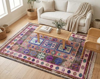 Könnte beinhalten: Ein rechteckiger Teppich mit einem verblassten, abgenutzten Muster und geometrischen Motiven in Rosa-, Lila-, Blau- und Brauntönen. Der Teppich hat einen hellen Rand mit kreisförmigen Akzenten und Fransen an den kurzen Seiten.
