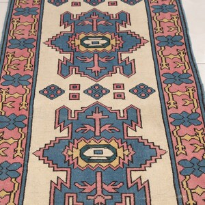 K&ouml;nnte beinhalten: Ein cremefarbener Teppich mit einem blauen, rosa und gelben geometrischen Muster. Der Teppich hat einen Rand mit einem sich wiederholenden floralen Muster.