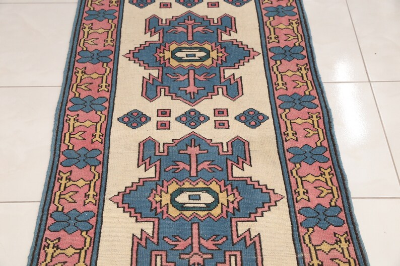 K&ouml;nnte beinhalten: Ein cremefarbener Teppich mit einem blauen, rosa und gelben geometrischen Muster. Der Teppich hat einen Rand mit einem sich wiederholenden floralen Muster.