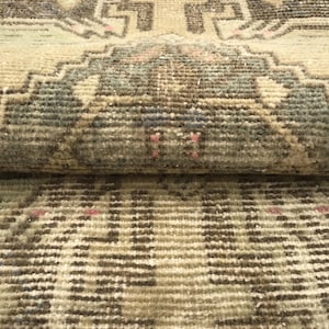 May include: A woven rug featuring a geometric pattern in shades of beige, brown, and muted green. The textile displays a folded section revealing a dense, knotted pile construction with small accents of pink thread integrated into the design.