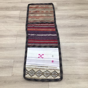 May include: A long, narrow rug with a black border and a multi-colored woven pattern. The rug is divided into four sections, each with a different pattern. The top section is a light brown and beige pattern, the second section is a red, purple, and white pattern, the third section is white with a pink cross and the bottom section is a brown and beige geometric pattern.