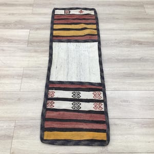 Könnte beinhalten: Ein langer, schmaler Teppich mit weißem Hintergrund und bunten geometrischen Mustern. Der Teppich hat einen schwarzen Rand und ist aus gewebtem Stoff.