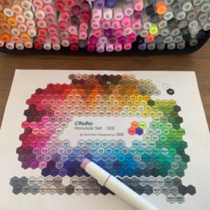 Pode incluir: Um conjunto de marcadores Ohuhu Honolulu Set - 320 em várias cores, dispostos numa bandeja. Um gráfico de cores com um padrão hexagonal é exibido, com um marcador em cima. O gráfico é projetado por Saar.