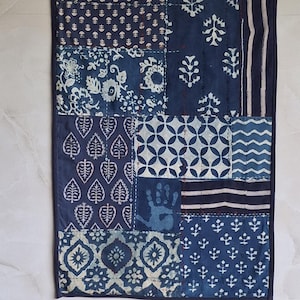 Puede incluir: Colgante de pared rectangular con diseño de patchwork en azul índigo. El tejido presenta varios patrones, incluyendo flores, hojas y formas geométricas. Los bordes están acabados con un borde azul oscuro.