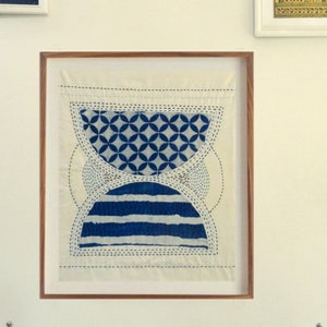 Puede incluir: Obra de arte textil enmarcada con un diseño geométrico. La obra presenta un fondo color crema con patrones azules, incluyendo círculos y rayas horizontales. La pieza está enmarcada en un marco de madera y montada en una pared blanca.