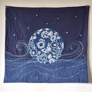 May include: A square textile wall hanging featuring a dark blue background with a central circular patch displaying a white floral pattern. The design includes white stitched wave motifs at the bottom and small white circular accents representing stars.