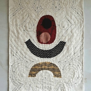 Könnte beinhalten: Abstraktes Textilkunstwerk mit cremefarbenem Hintergrund und geometrischen Formen. Es zeigt ein rotes Oval mit einem schwarzen Kreis, eine schwarz-weiße Zickzackform und ein braunes Bogendesign. Das Stück ist mit schwarzem Faden handgenäht.