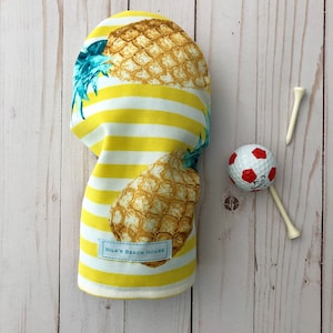 May include: Yellow and white striped golf headcover with a pineapple print and the text "Mila's Beach House".