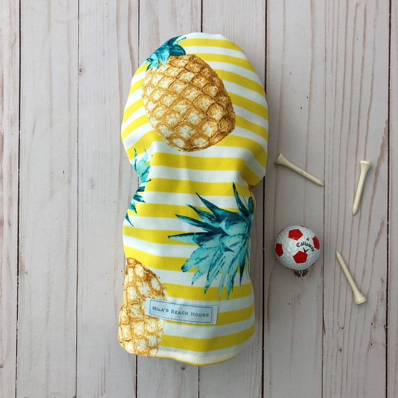 May include: Yellow and white striped golf headcover with a pineapple print. The headcover has the text "Mila's Beach House" printed on it.