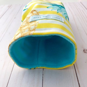May include: A yellow and white striped fabric tube with a blue fleece lining. The fabric has a floral pattern and the text "Mila's Beach House" is printed on the tube.