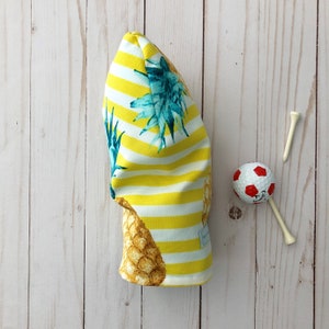 May include: A yellow and white striped golf headcover with a pineapple print. The headcover has a white tag with black text.