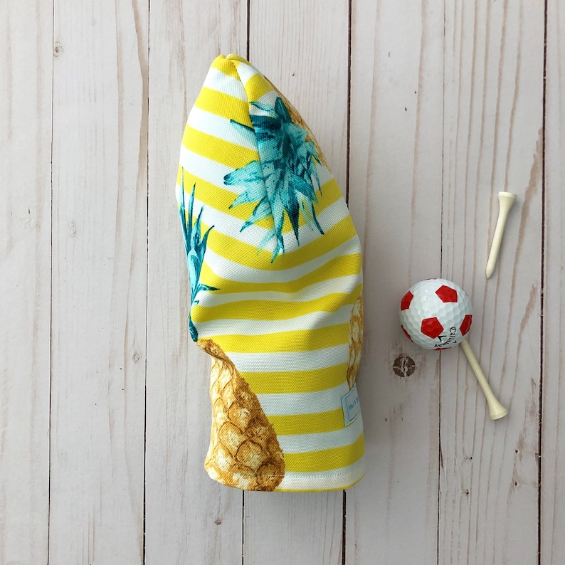 May include: A yellow and white striped golf headcover with a pineapple print. The headcover has a white tag with black text.