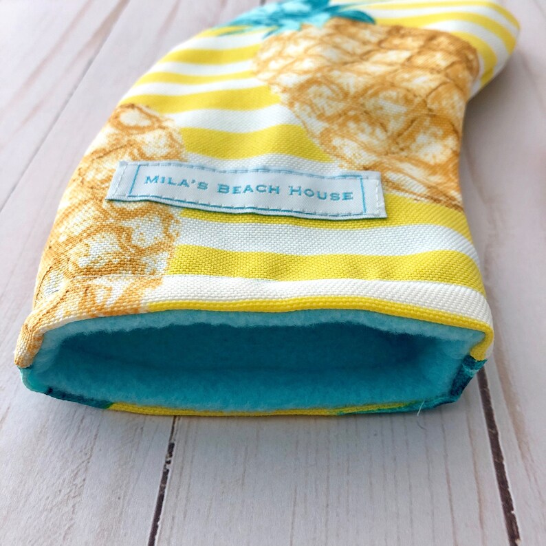 May include: A yellow and white striped fabric sleeve with a pineapple print and a blue fleece lining. The sleeve has a white label with the text "Mila's Beach House".