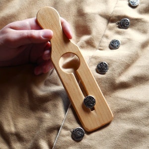 May include: A light-colored wooden button hook tool with a handle and a keyhole-shaped opening. The tool is held over a tan fabric with decorative silver buttons.