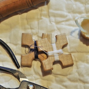 May include: Three wooden cross-shaped weaving tools with blue and white thread wrapped around them. A pair of metal shears and a wooden tool are also visible. The items are on a cream-colored, quilted surface.
