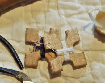 May include: Three wooden cross-shaped weaving tools with blue and white thread wrapped around them. A pair of metal shears and a wooden tool are also visible. The items are on a cream-colored, quilted surface.