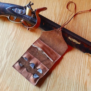 May include: A brown leather pouch with several compartments, holding flint stones and a small wooden rod. A vintage firearm with a dark wooden stock and metal components is partially visible in the background. The pouch is open, displaying the contents.