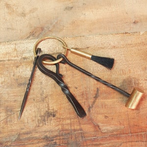 May include: A collection of black metal tools attached to a brass ring, including a pick, a hook, and a small hammer with a brass head. A small brush with black bristles is also included. The tools are arranged on a wooden surface.