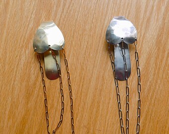 May include: Two silver-toned metal pendants with a heart-shaped top and a long, chain-like bottom. The pendants are displayed on a light brown wooden surface. The chains are attached to the bottom of the heart-shaped top.