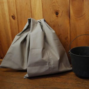 May include: A large, gray canvas bag sits on a wooden floor next to a black metal bucket.