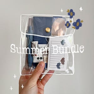 May include: A clear plastic pouch labeled "Summer Bundle" containing a navy blue notebook, pen, and other stationery items. The pouch is held by a hand, with decorative blue flowers and stars in the background.