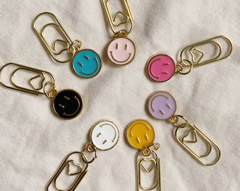 May include: Set of eight gold paper clips with colorful enamel smiley face charms. Each clip features a heart-shaped opening.