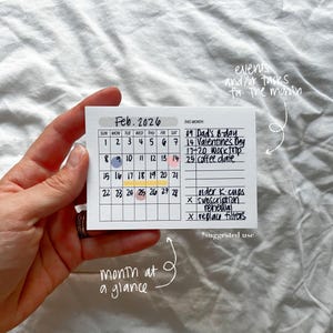May include: A small, rectangular calendar for February 2026, held in a hand. The calendar features a grid layout with dates and space for notes. Key dates like Valentine's Day and Dad's birthday are highlighted. The text "month at a glance" is also visible.