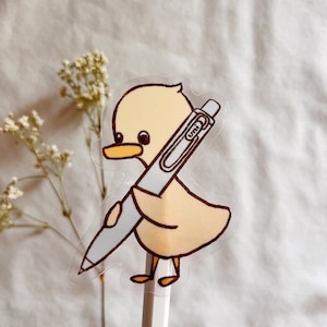 May include: A clear sticker of a yellow duckling holding a pen with the word "UNI" on it. The sticker is attached to a white pencil.