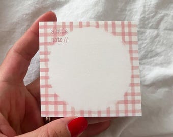 Peut inclure: Un bloc-notes carré avec un motif vichy rose et blanc et un grand espace blanc circulaire au centre pour écrire. Le texte 'a little note //' est imprimé dans le coin supérieur gauche dans une police sans empattement simple et foncée.