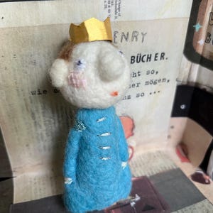 Può includere: Una bambola di feltro che indossa un cappotto blu con bottoni argentati e una corona dorata. La bambola ha un viso bianco con capelli castani e guance rosa.
